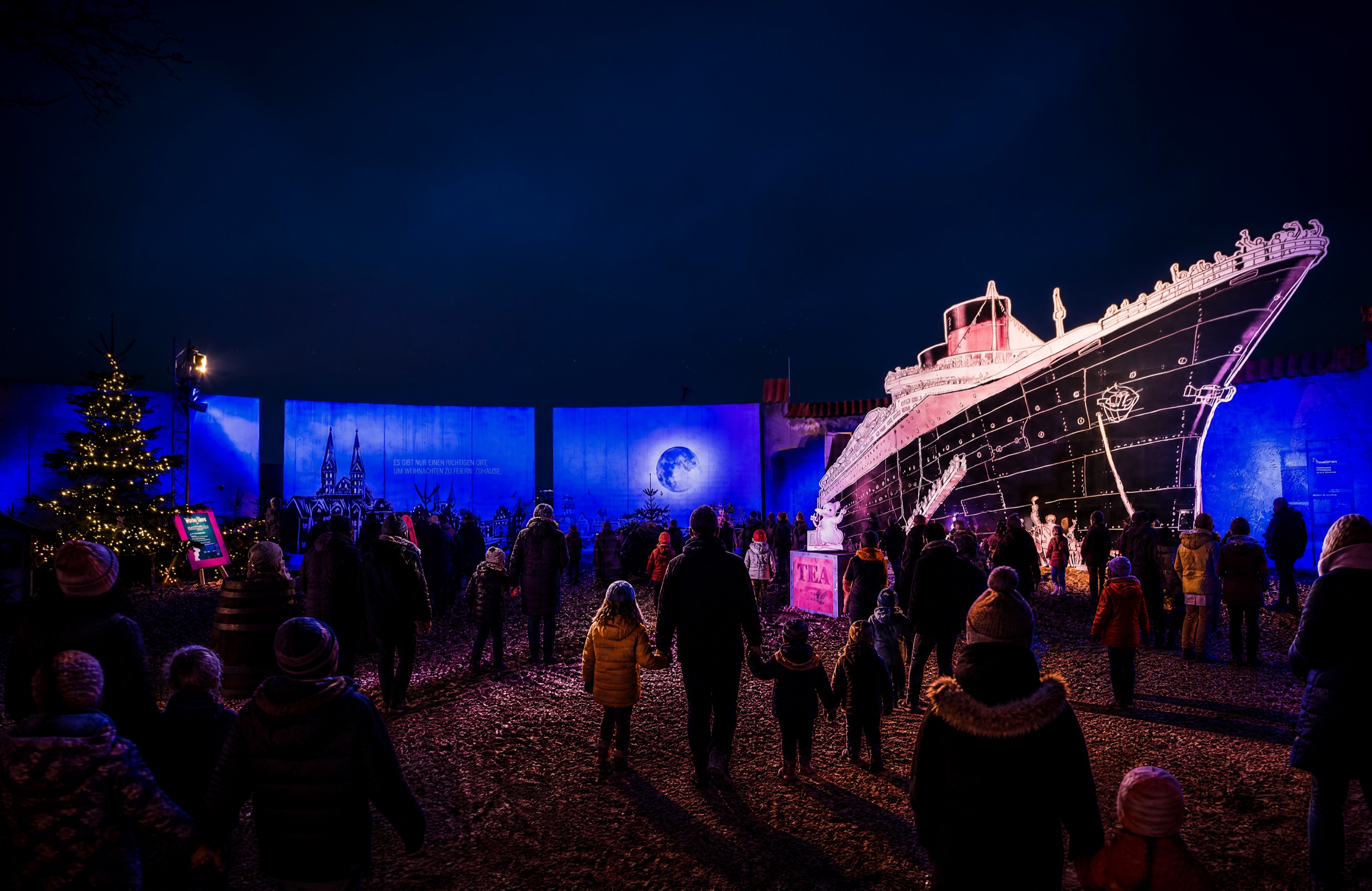 Families and visitors gather around the illuminated Hanseatic ship in front of the blue panoramic wall at the European Hansemuseum.